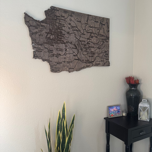 Wooden wall art of a map of Washington state on a white wall with a plant and small table in the foreground.
